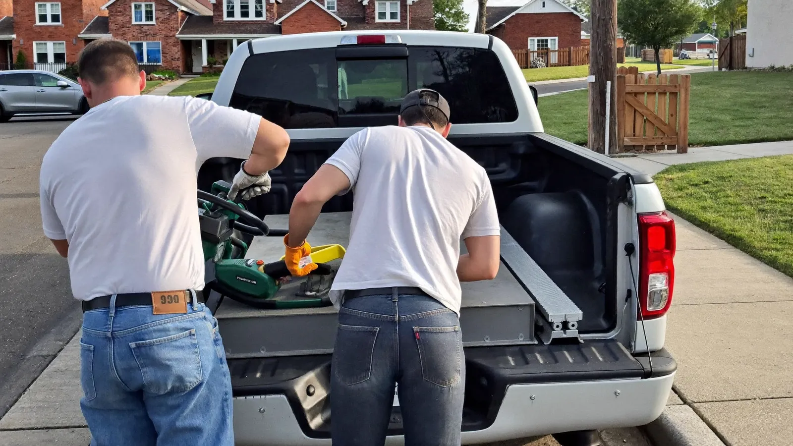 Two foundation technicians in work clothing beside a pickup truck loaded with pier sections on a residential street of brick homes in Edmond, Oklahoma